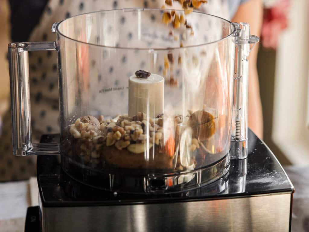 A food processor is filled with various nuts and vegan sausage and garlic, which are falling from above. A person stands blurred in the background wearing a polka dot shirt.